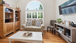Un salon chaleureux avec un meuble en bois, un canapé agrémenté de coussins ornés de la mention « VIVEZ, AIMEZ, PROFITEZ DU LAC », une table basse et un téléviseur fixé au mur.