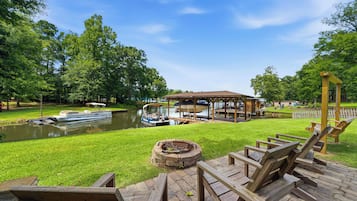 Une terrasse au bord du lac, avec des chaises en bois, un foyer et un belvédère offrant une vue sur un lac paisible où naviguent des bateaux.