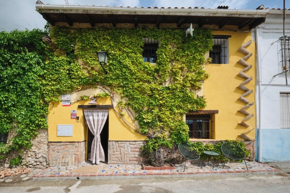 A yellow building with a stone entrance, a door with a curtain, and a small outdoor seating area with two chairs.