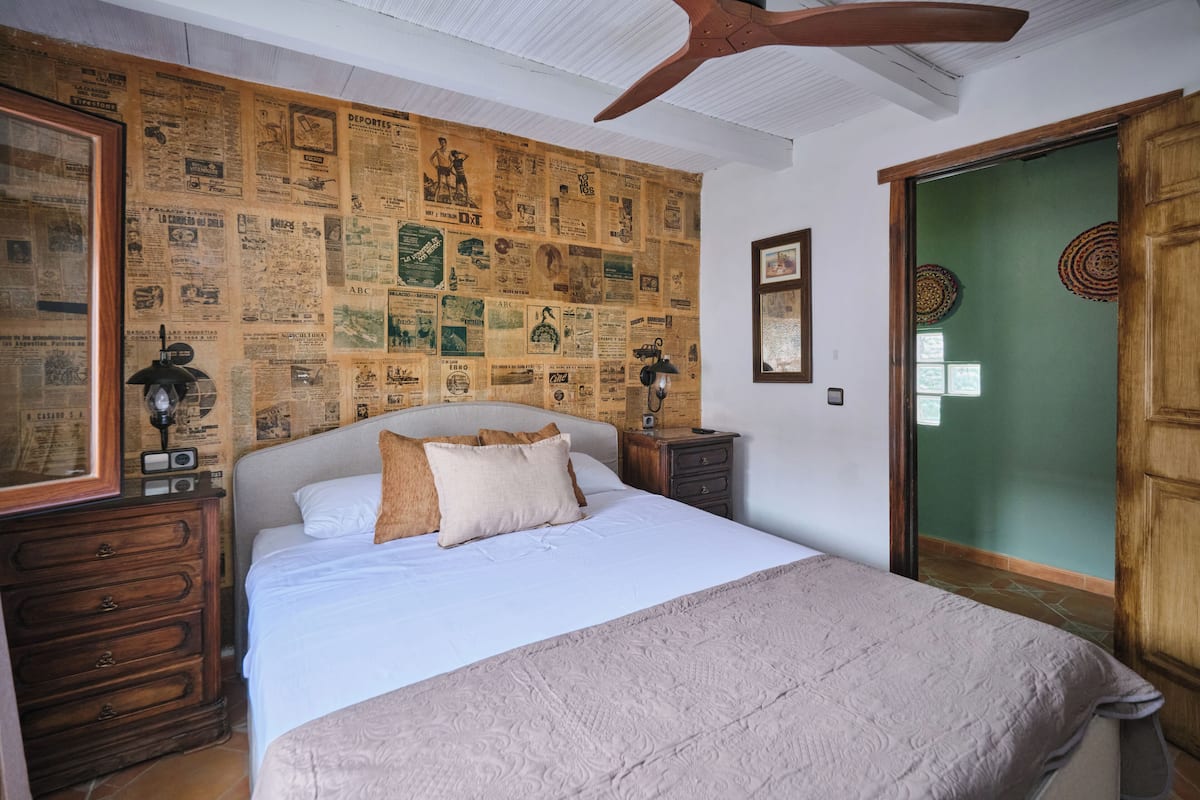 A bedroom with a large bed, wooden headboard, a ceiling fan, a wall covered with newspaper clippings, and a green door.