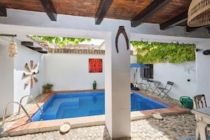 A swimming pool with a tiled edge, a wooden deck, a table with chairs, and a wall with a decorative pattern. - Casa Mathilda (Fornes)