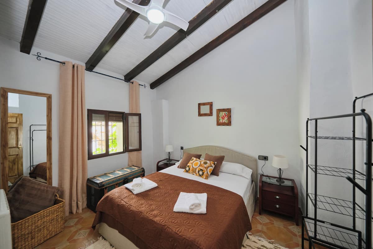 A bedroom with a bed, a nightstand, a lamp, a mirror, a window, and a ceiling fan.