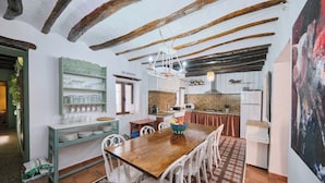 A dining area with a wooden table, white chairs, a green sideboard with glassware, and a kitchen in the background. - Casa Mathilda (Fornes)
