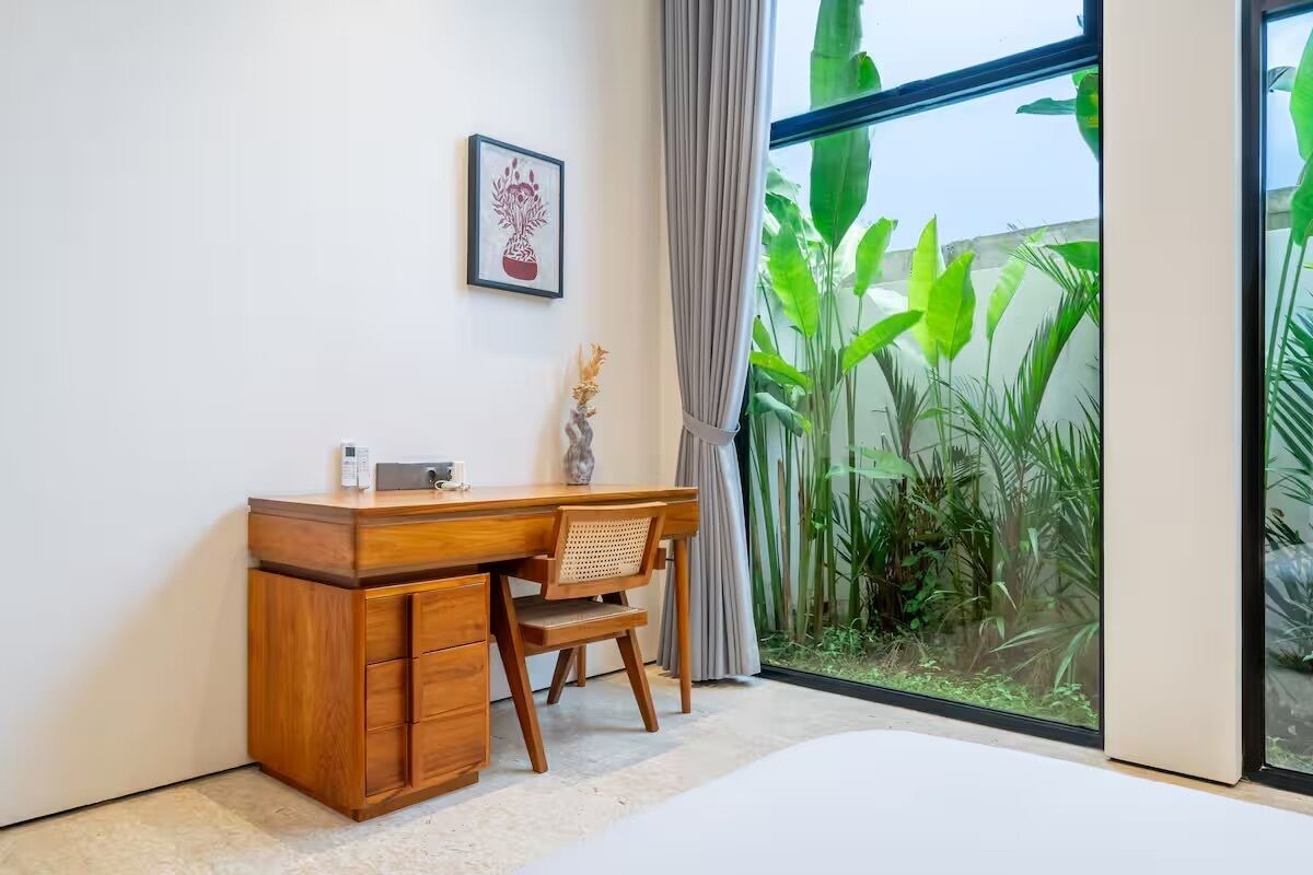 A room with a wooden desk, a chair, a bed, a window with a view of greenery, and a framed picture on the wall.