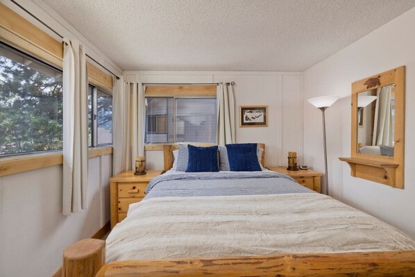 A bedroom with a wooden bed, two bedside tables, a window with curtains, a bedside lamp, a mirror, and a framed picture on the wall. - Sierra Manors Mountain Escape (Mammoth Lakes)