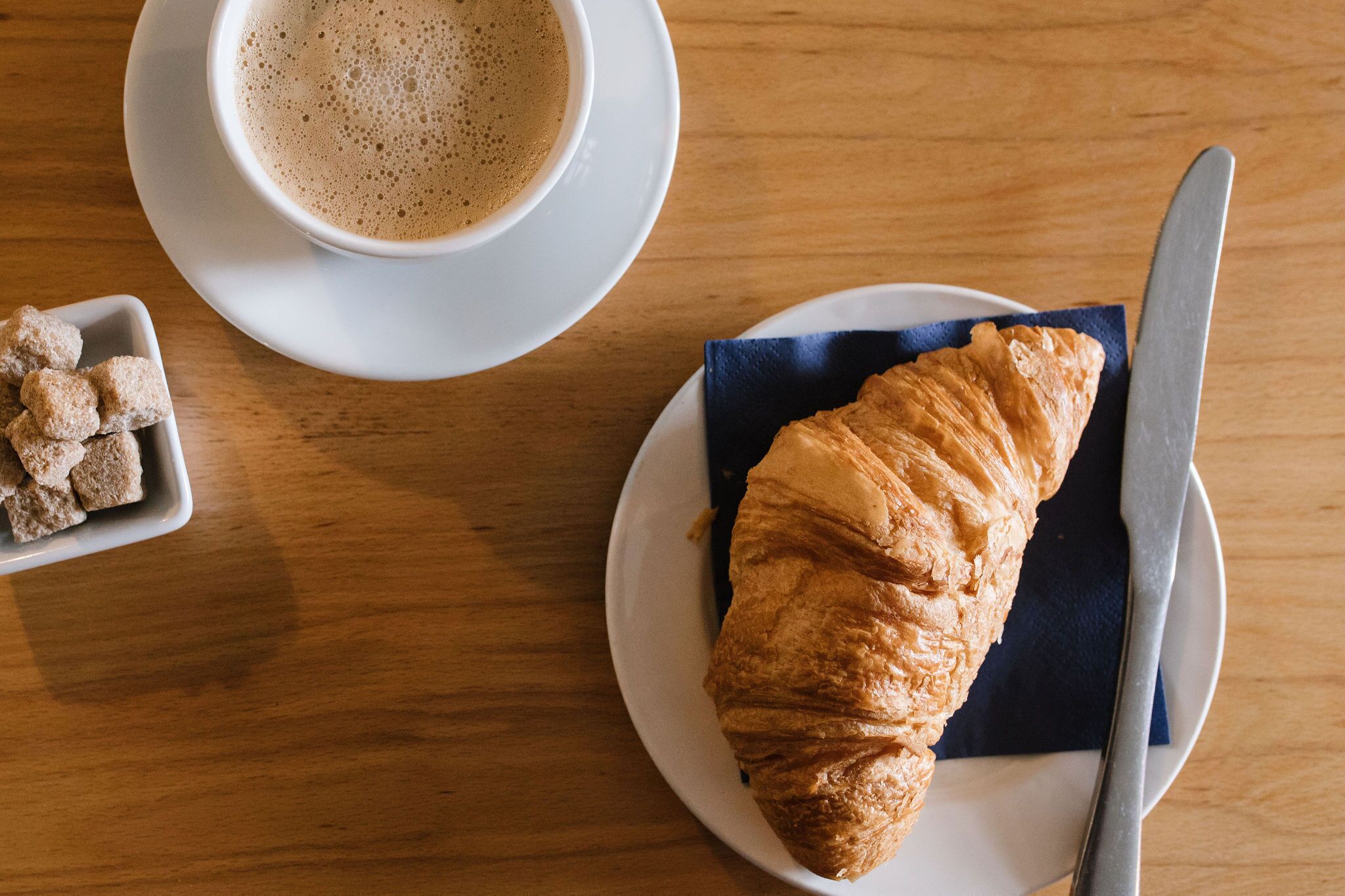 Un croissant sur une assiette, accompagné d’un couteau, une tasse de café et un petit bol de morceaux de sucre, le tout posé sur une table en bois.