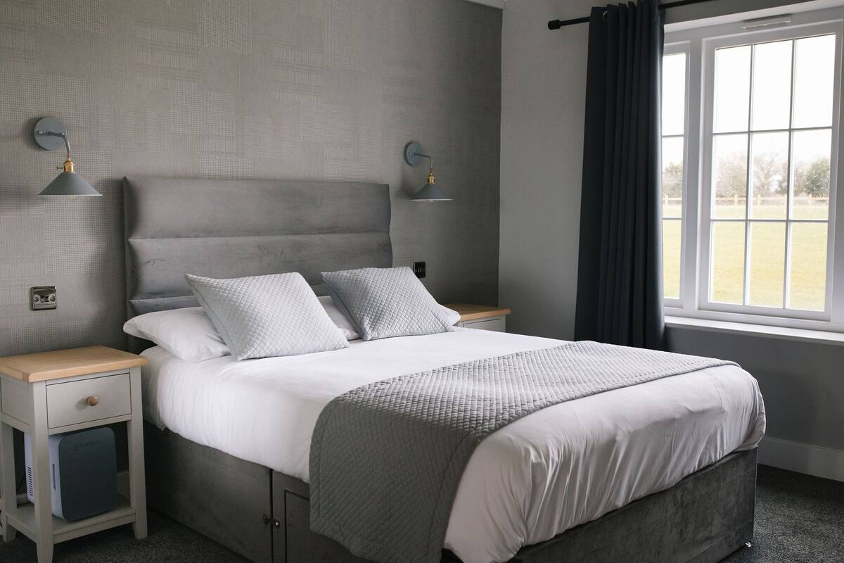 Un dormitorio con una cama, dos mesitas de noche, un velador y una ventana con cortinas, con vistas al exterior.