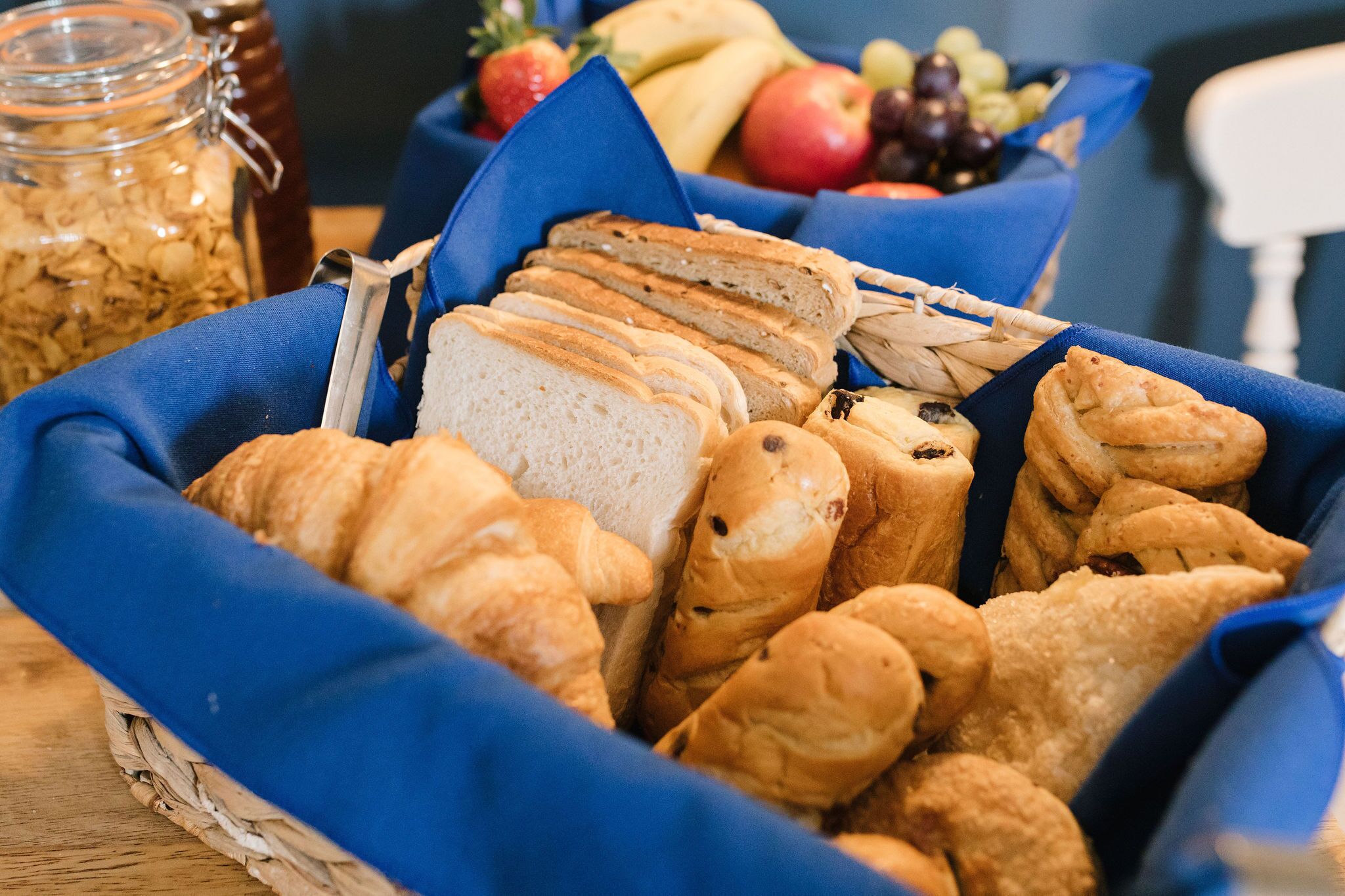 A basket of assorted breads and pastries, a jar of granola, and a bowl of fruit.