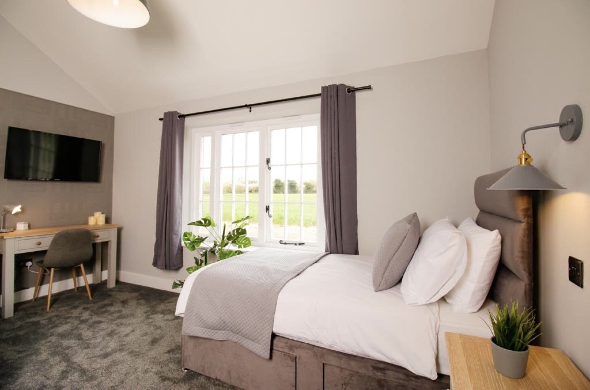 Une chambre à coucher comprenant un lit, un bureau, une chaise, une télévision et une fenêtre avec des rideaux.