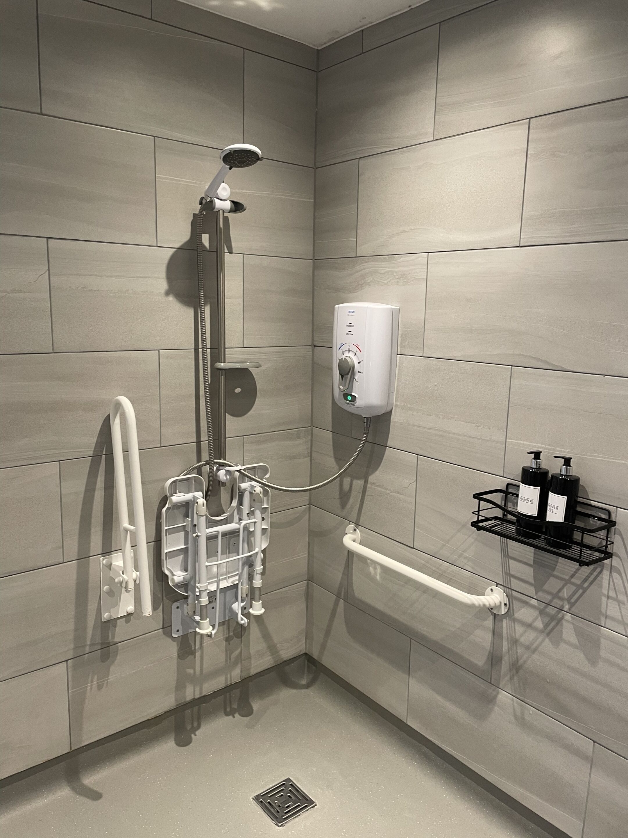 A modern shower with a handheld showerhead, a wall-mounted soap dispenser, and a handrail.