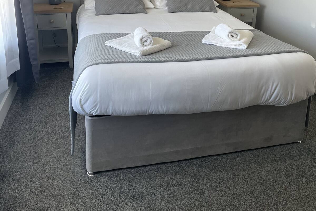 Una cama bien hecha con dos almohadas, una mesita de noche con una lámpara y un cajón pequeño.
