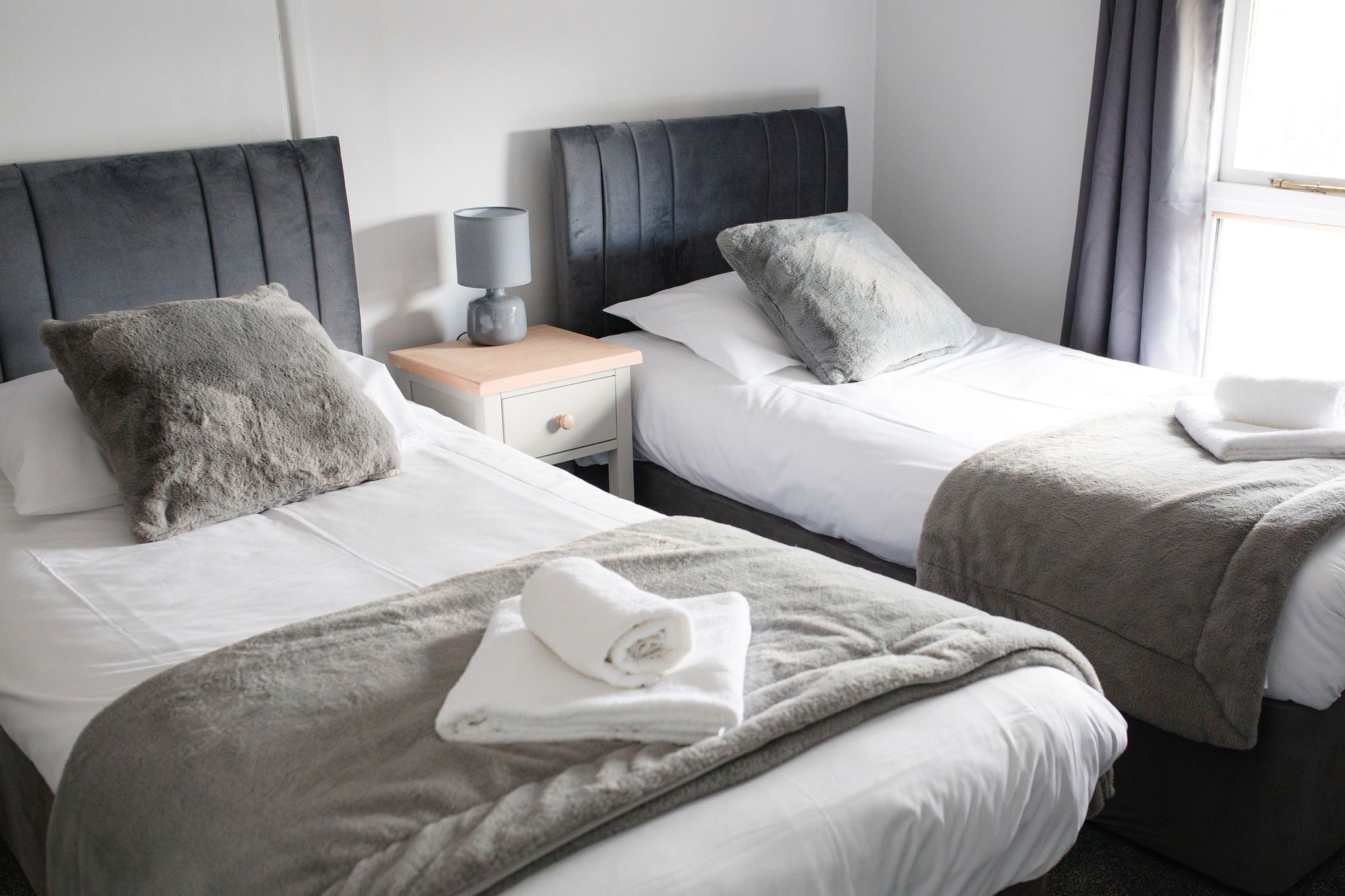 Habitación de hotel con dos camas, una mesita de noche con lámpara y una ventana con cortinas.