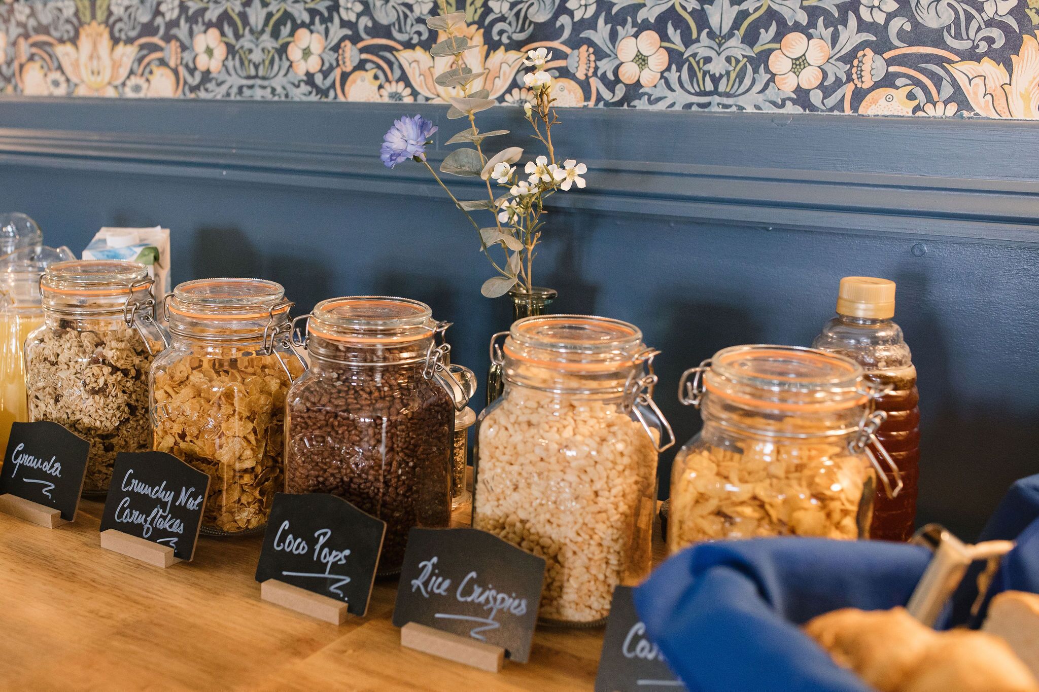 A display of various food items in glass jars with labels, set against a patterned wall.