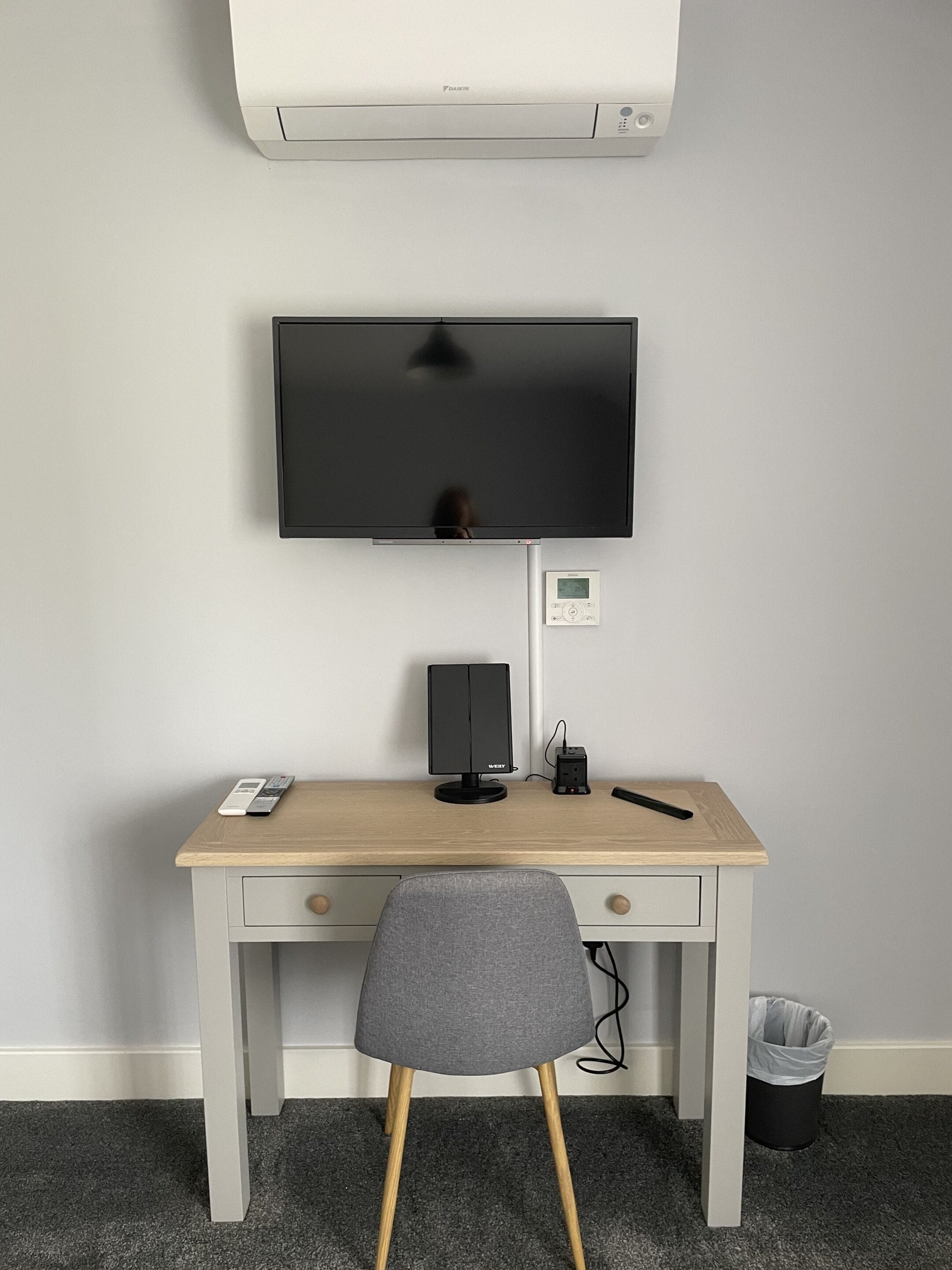 A room with a desk, a television mounted on the wall, a speaker, a remote control, and a trash bin.