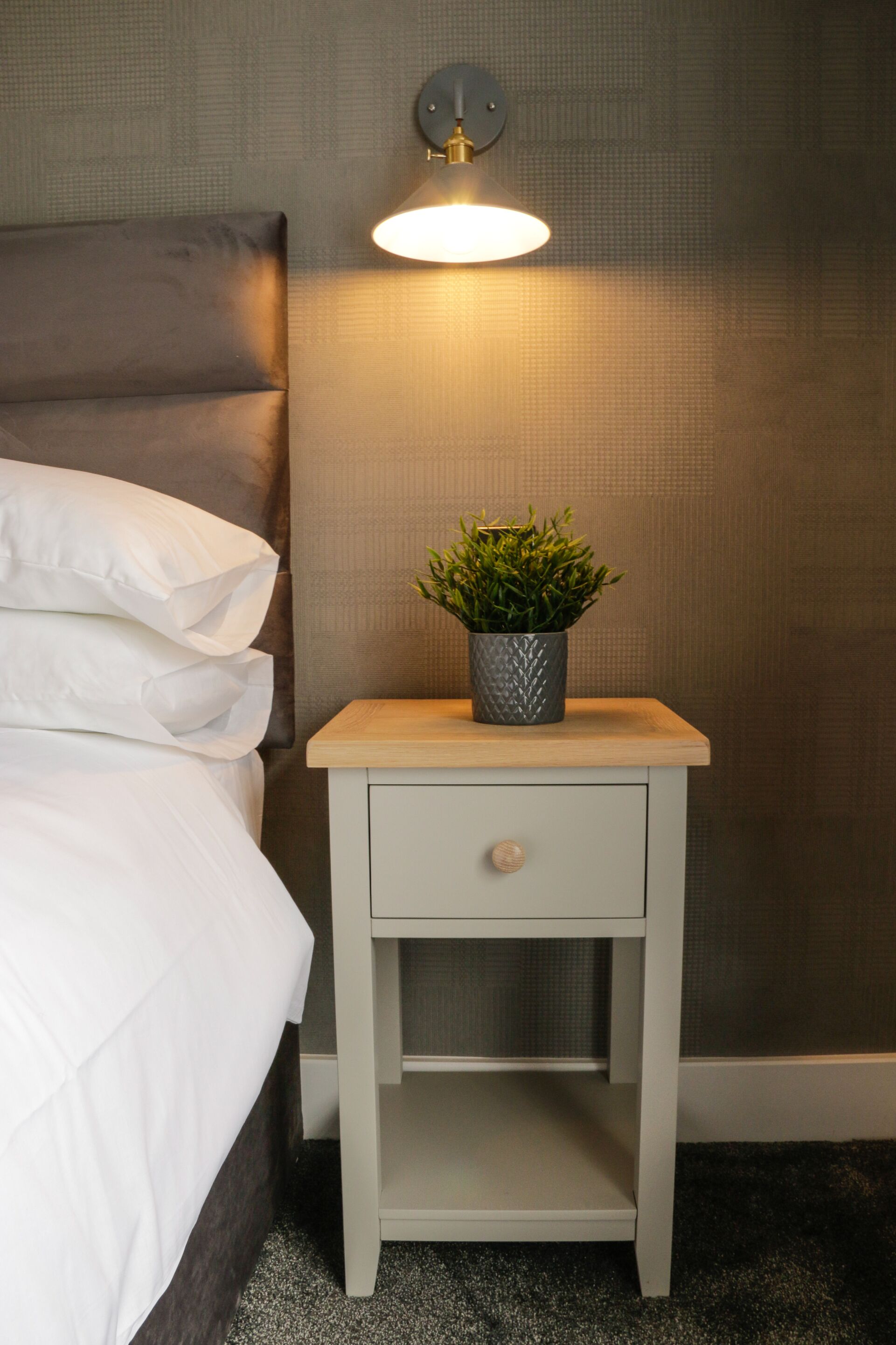 Ein ordentlich bezogenes Bett mit weißen Leinentextilien, ein kleiner Nachttisch mit einer Topfpflanze und eine Wandlampe.