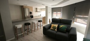 A modern kitchen with a built-in oven and microwave, a bar area with stools, a dark sofa with a leaf-patterned pillow, and a large window providing natural light. - Golden Sky Big Families (Benicarló)