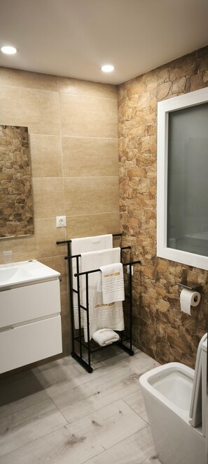 A bathroom with a stone wall, a white sink, a black towel rack with towels, and a toilet. - Golden Sky Big Families (Benicarló)