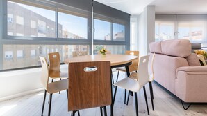 Una sala de estar moderna con una mesa redonda de madera, sillas color beige, un sofá color beige y ventanas grandes que ofrecen vistas a edificios.