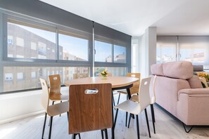 Una sala de estar moderna con una mesa redonda de madera, sillas color beige, un sofá color beige y ventanas grandes que ofrecen vistas a edificios.