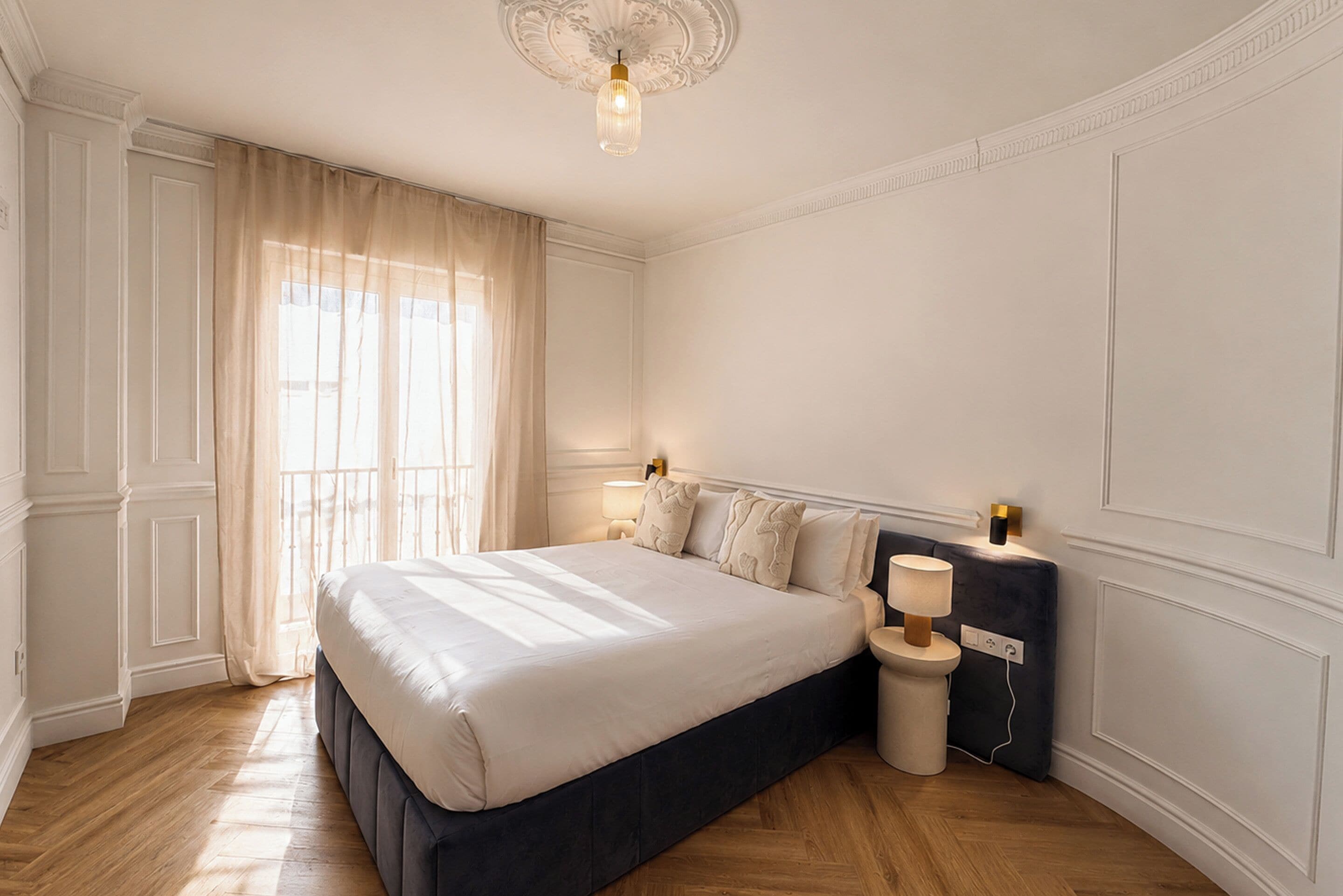 A bedroom with a large bed, a nightstand, a bedside table, a wall-mounted lamp, and a window with curtains.