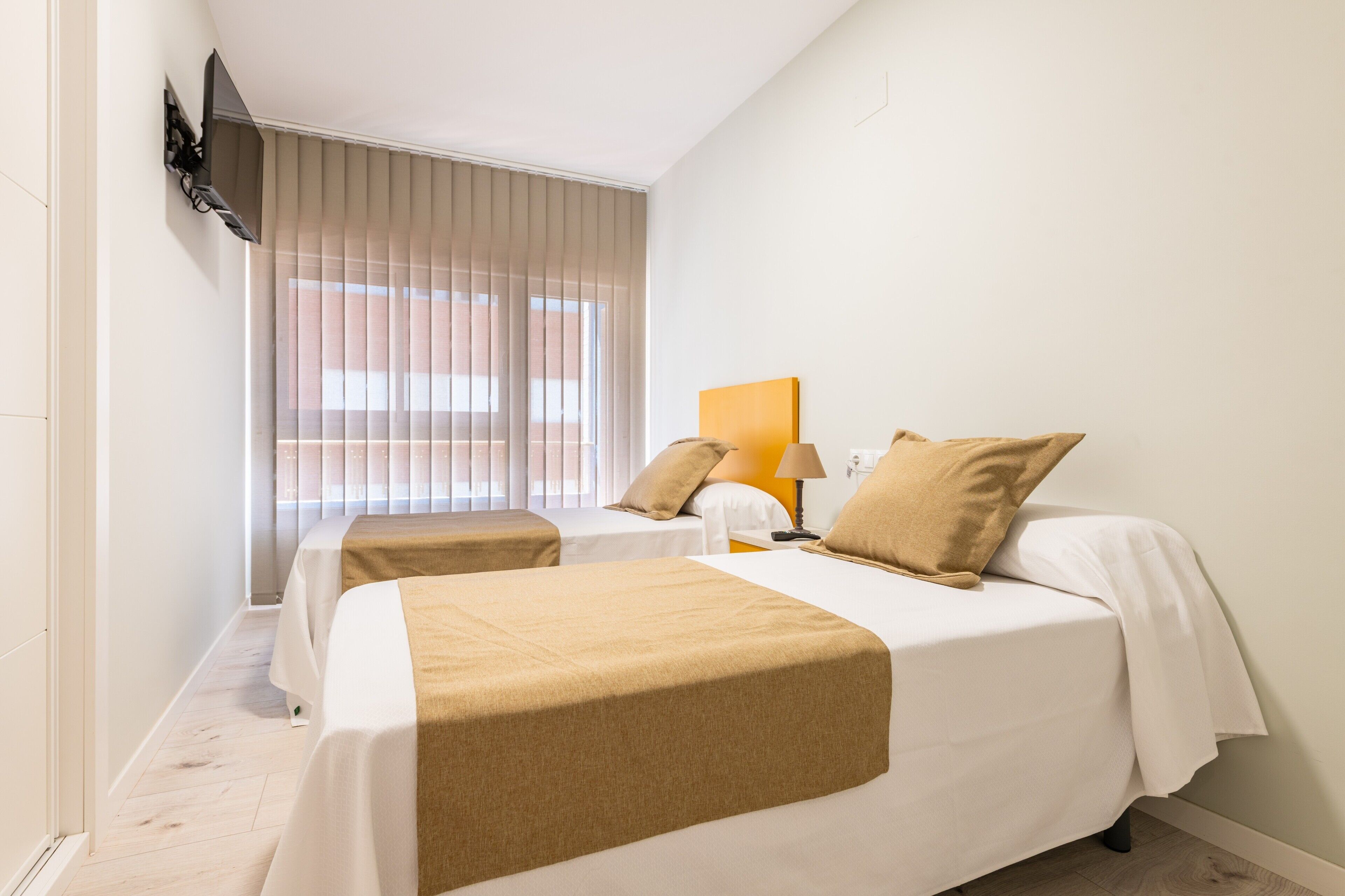 Ein Hotelzimmer mit zwei Betten, einem an der Wand befestigten Fernseher und einem Fenster mit Jalousien.