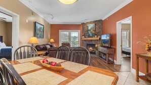 Een eetgedeelte met een houten tafel voor vier personen, een open haard, en een woonkamer met een bank en televisie.