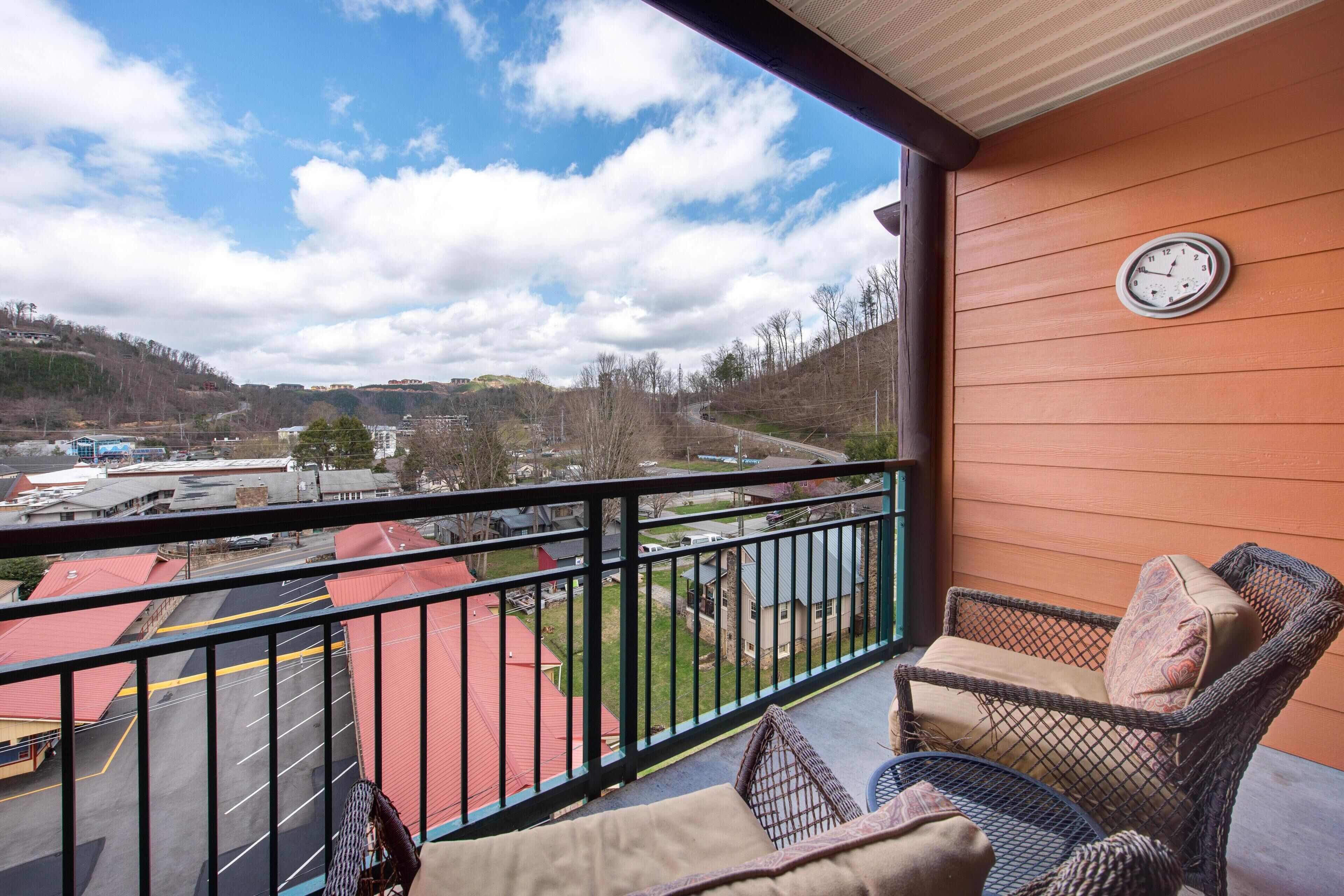 Un balcon donnant sur un parking et des immeubles, une chaise en osier avec des coussins et une horloge fixée au mur.