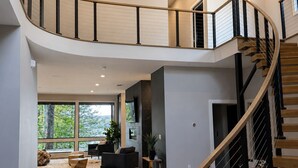 A spacious interior with a curved staircase, wooden flooring, and large windows offering a view of greenery. - Modern Deep Creek Lakefront Home, Movie Theater, Hot Tub, 7BR/6BA, Sleeps 16 (Oakland)