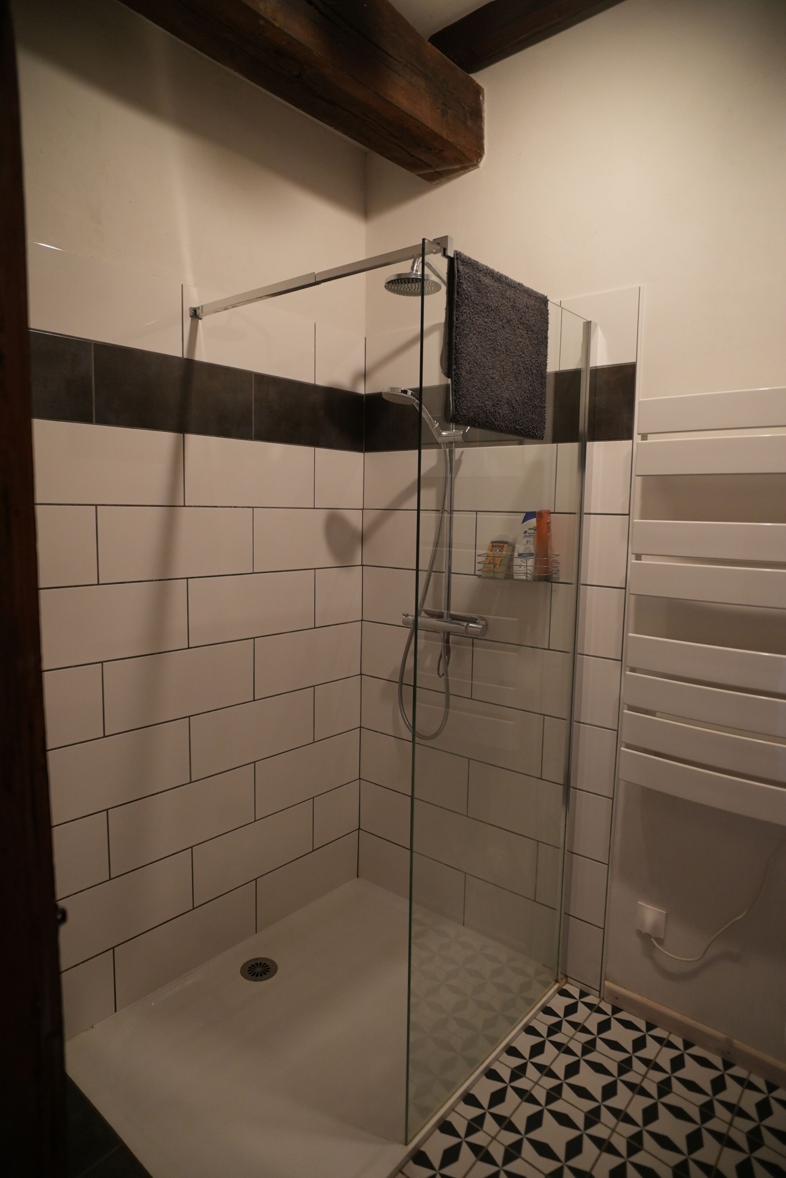 A shower with a glass door, white tiled walls, a black and white patterned floor, and a wooden beam above.