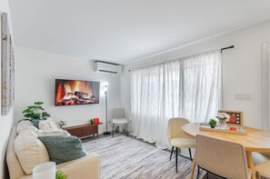 Ein modernes Wohnzimmer mit einer Couch, einem Esstisch mit Stühlen, einem Gemälde eines Feuers und einem großen Fenster mit Vorhängen.