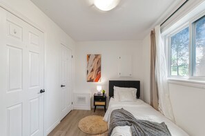 Ein Schlafzimmer mit Bett, Nachttisch, Wandbild, Fenster mit Vorhängen und einer Tür.