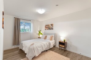 Ein Schlafzimmer mit Bett, Nachttisch, Lampe und Fenster mit Vorhängen.