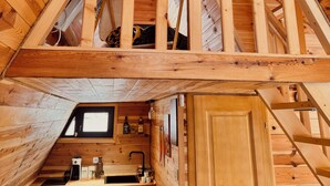 Interior de uma cabana de madeira com quarto no mezanino, área de cozinha e uma pequena janela.