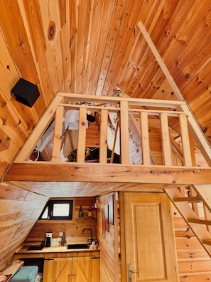 A wooden cabin interior with a loft bedroom, kitchen area, and a small window.