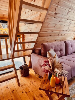 Un intérieur en bois avec un canapé, une table ornée d’un vase et de fruits, et une fenêtre donnant sur un paysage.