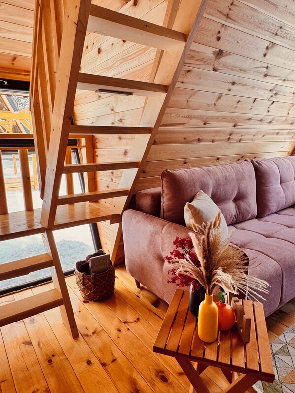 Un intérieur en bois avec un canapé, une table ornée d’un vase et de fruits, et une fenêtre donnant sur un paysage.