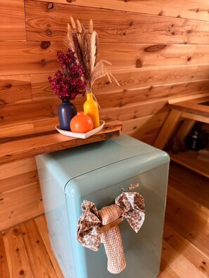 Une pièce aux murs en bois, une petite table sur laquelle est posé un vase de fleurs, et une valise bleu clair ornée d’un ruban.