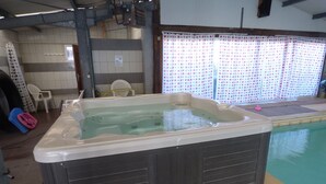A hot tub in a room with tiled walls, a window with curtains, and a pool outside.