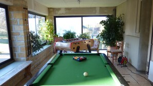 Una mesa de billar con superficie de fieltro verde, una bola blanca y una mesa de fútbol de mesa al fondo.