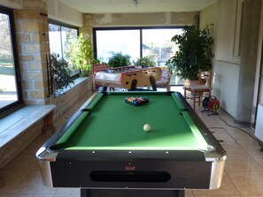 A pool table with a green felt surface, a single white cue ball, and a foosball table in the background.