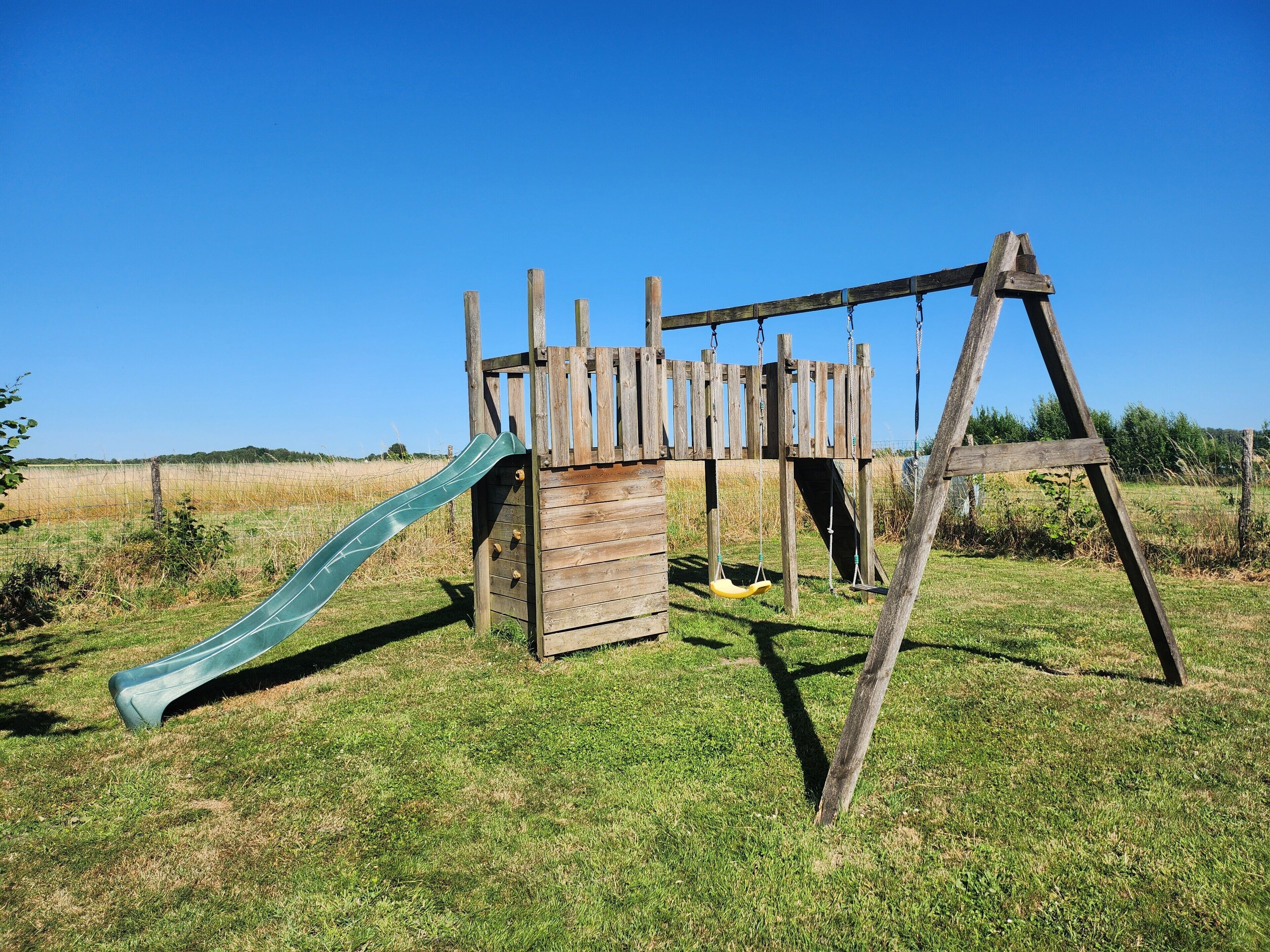Een houten speeltoestel met een glijbaan en schommels in een grasveld.
