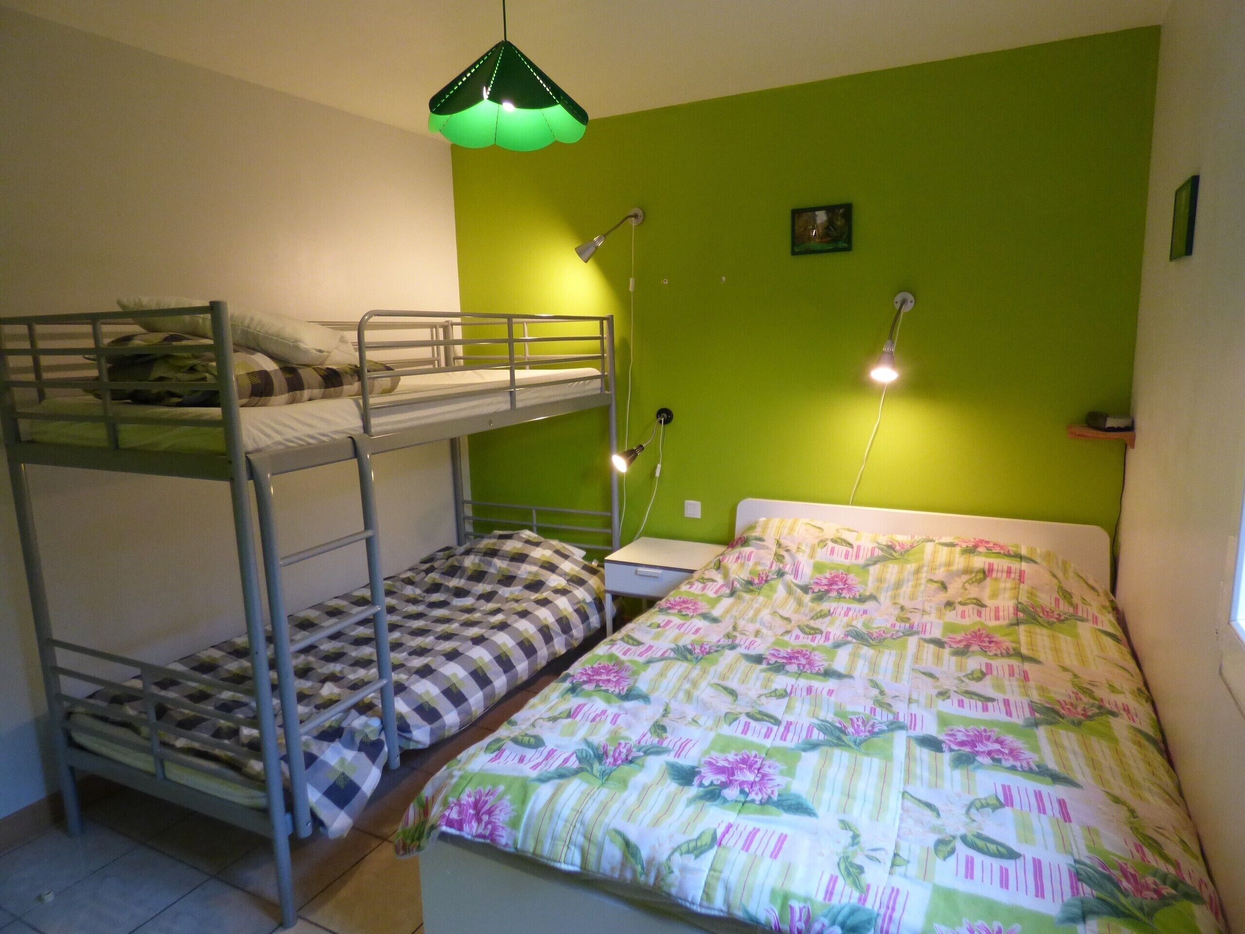 A bunk bed room with a green wall, a single bed with floral bedding, and a desk with a lamp.