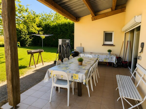 Une terrasse extérieure avec une table dressée pour le repas, entourée de chaises, et un barbecue.