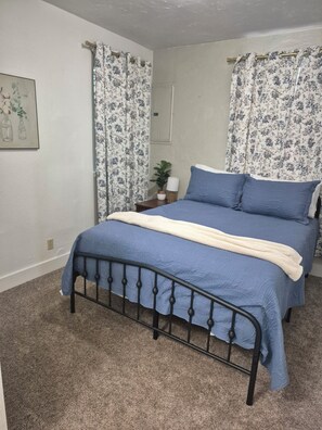 A bedroom with a bed, curtains, a nightstand, a lamp, and a wall-mounted mirror.