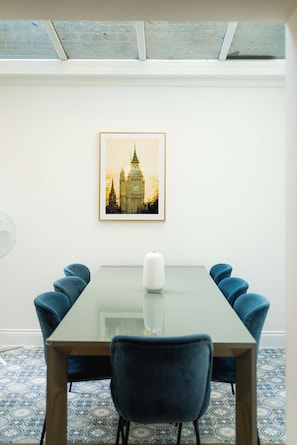 Un comedor con una mesa de vidrio, sillas tapizadas en azul, un cuadro enmarco de un edificio y baldosas de piso estampadas.