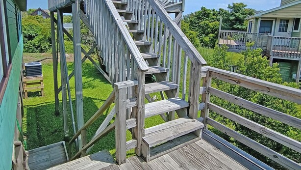 Un escalier en bois mène à une terrasse dotée d’une rambarde, le tout entouré de verdure et surplombé par un ciel dégagé.