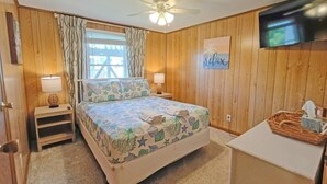 Un dormitorio con cama, ventilador de techo, ventana con cortinas, televisor montado en la pared y mesita de noche con lámpara.