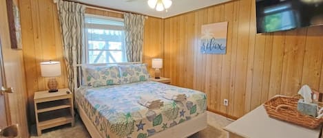 A bedroom with a bed, a ceiling fan, a window with curtains, a wall-mounted TV, and a bedside table with a lamp.