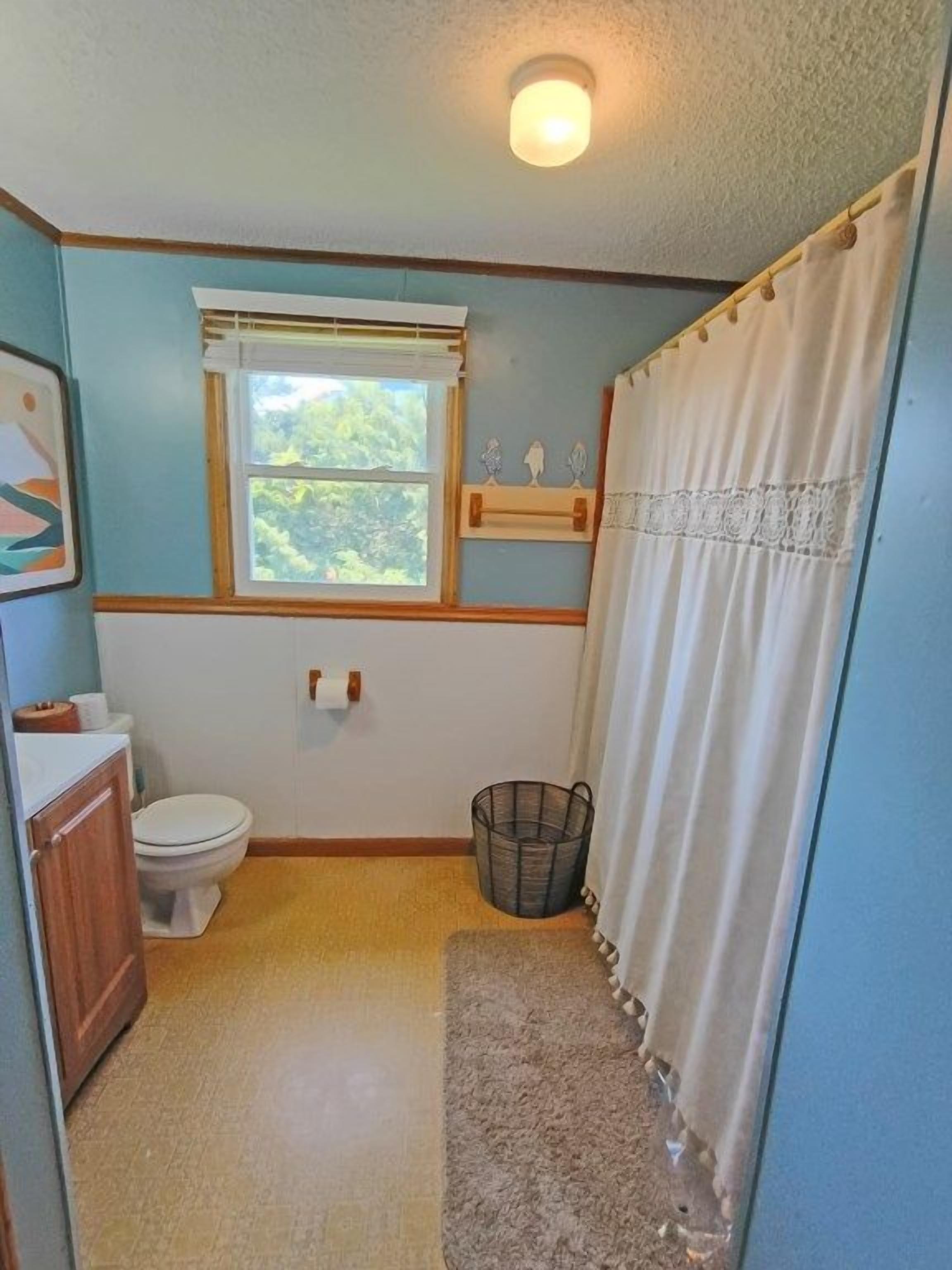 A small bathroom with a toilet, a window, a shower curtain, and a wooden cabinet.