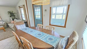 Un coin repas avec une table en bois, des chaises en osier, un chemin de table à motifs et une fenêtre avec des stores.